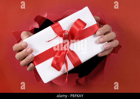 Weiß Geschenk mit Schleife durch ein rotes zerrissenes Papier Hintergrund geliefert wird. Stockfoto