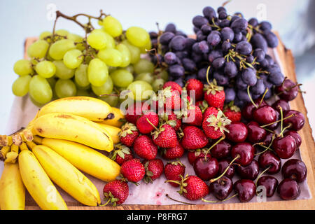 Früchte (weiße und blaue Trauben, Bananen, Erdbeeren und Kirschen) auf einer Holzplatte Stockfoto