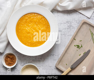 Eine Schale hausgemachter Kürbissuppe neben kleinen Schüsseln mit Muskatnuss und Sahne und Schnittlauch auf einer hölzernen Schneidebrett. Stockfoto