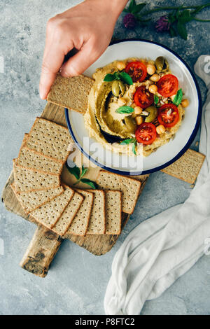 Eine Frau, die ein Cracker eintauchen in eine Schüssel mit Hummus ohne tahini mit Cherry Tomaten und grüne Oliven gekrönt. Stockfoto