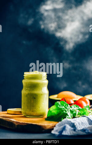 Avocado Dressing in einem Glas auf einem Holzbrett von vorne fotografiert. Rucola Blätter, Tomaten, Avocado mit seiner Grube begleiten. Stockfoto