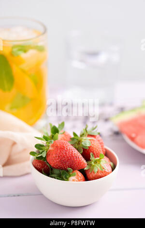 Nahaufnahme von einem weißen Keramik Schüssel mit Erdbeeren auf den Tisch und ein Glas Glas mit Eistee im Hintergrund Stockfoto