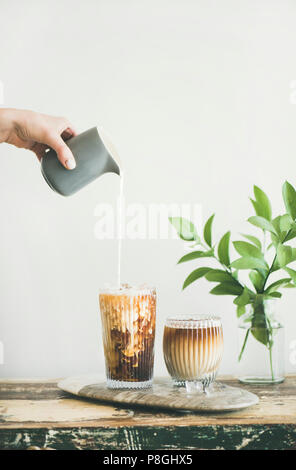 Eiskaffee in hohe Gläser mit Milch gegossen von Krug von Hand, weiße Wand und grünen Zweigen in Vase im Hintergrund, kopieren. Sommer Stockfoto