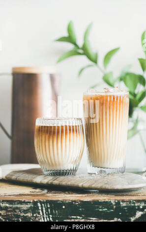 Eiskaffee in hohe Gläser mit Milch an Bord, weiße Wand, grüne Pflanze Niederlassungen und Kanne im Hintergrund, Kopieren, close-up. Sommer erfrischende bevera Stockfoto