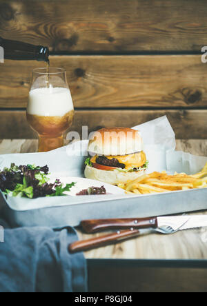 Classic Burger essen. Rindfleisch hausgemachten Burger mit Glas Lagerbier, Pommes frites und Salat auf weißen Fach, rustikalen Holzmöbeln Wand im Hintergrund, cop Stockfoto