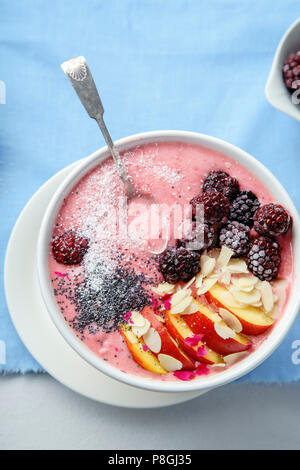 Frischen und kalten strawberry Banana Smoothie garniert mit peach Slices, gefroren Blackberry, Chia Samen und Mandelflocken. Blau Leinen Hintergrund Stockfoto