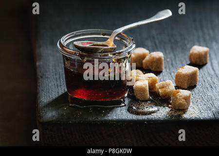 Hausgemachte Flüssigkeit transparent Brown Sugar caramel im Glas steht auf schwarze Holzbrett mit Löffel und können Würfel Zucker. Close Up. Tag Licht