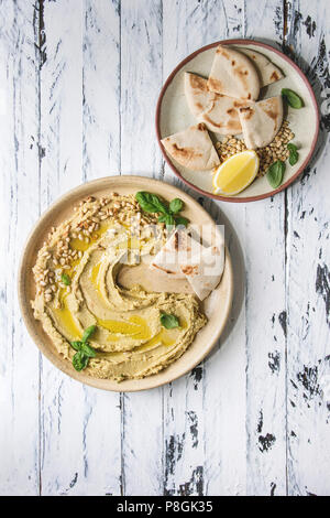 Hausgemachte, traditionelle Verbreitung Hummus mit Pinienkernen, Olivenöl, Basilikum, serviert auf Keramikteller mit pita Brot über weiß Holzbrett Hintergrund. Flat l Stockfoto