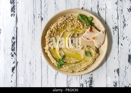 Hausgemachte, traditionelle Verbreitung Hummus mit Pinienkernen, Olivenöl, Basilikum, serviert auf Keramikteller mit pita Brot über weiß Holzbrett Hintergrund. Flat l Stockfoto