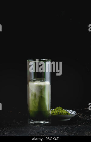Matcha Grüntee Iced Latte oder Cocktail in hohes Glas mit Eiswürfeln und Matcha Pulver über dunkle Textur Hintergrund Stockfoto