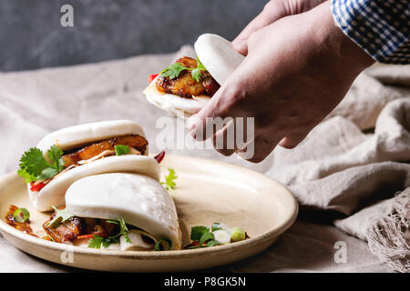 Des Menschen Hände halten asiatische Sandwich gedämpft Gua bao Brötchen mit Schweinebauch, grünen und Gemüse in Keramik Teller am Tisch serviert mit Bettwäsche Tischdecke. Wie Stockfoto