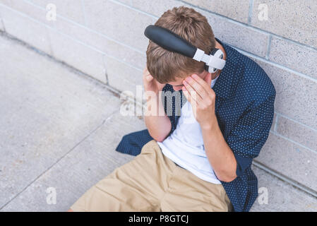Mit Kopfschmerzen Musik hören, während auf dem Boden sitzend gegen eine Wand jugendlich. Stockfoto