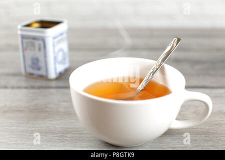 Heiße Tasse gelb Tee mit Dampf, mit Metall Zinn auf grau Holz Schreibtisch im Hintergrund platziert. Stockfoto