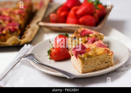 Erdbeeren und Rhabarber Torte serviert auf schöne Platte mit Holz- Schüssel mit Erdbeeren und ganze Backblech der Torte im Hintergrund Stockfoto