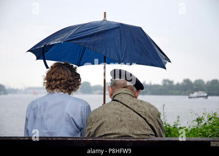 Ein Senioren-Paar auf einer Parkbank an der Promenade in Hamburg-Teufelsbrueck schuetzt sich mit einem Regenschirm gegen einen Sommer-Regen. (C) Marti Stockfoto