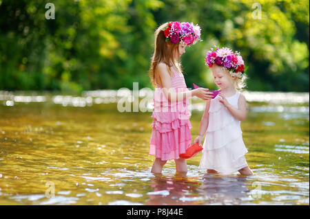 Zwei Entzückende kleine Schwestern tragen Blume Kronen spielen mit Papier Boote durch einen Fluss an warmen und sonnigen Sommer Tag Stockfoto