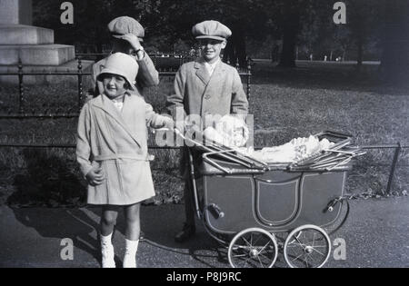 1920s, historische, drei elegant gekleidete Kinder, die von einem Park das Tragen der Kleidung des Tages - die beiden Jungs haben beide flacher Deckel Hüte auf", newsboy' mit Stil - mit den jungen Mädchen Stolz von pram ihre hübschen Puppen, England, UK. Stockfoto