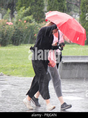 Belgrad, Serbien - Juni 14, 2018: Zwei junge Frauen unter roten Regenschirm in die plötzliche schwere Feder Regen in der Stadt Park läuft, Halten einer Kamera und Stockfoto