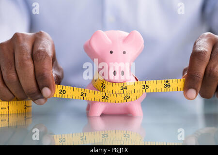 Einer Person Hand Messen rosa Sparschwein mit Maßband auf reflektierenden Schreibtisch Stockfoto