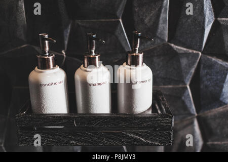 Drei weiße Metall Flaschen Shampoo, Spülung, Duschgel auf Holzregal im modernen Hotel Bad mit schwarzen Strukturlack Wand Stockfoto