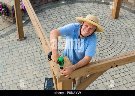 Gerne zuversichtlich Mann in einem strohhut Errichtung einer neuen Pavillon im Freien auf einem Stein gepflasterte Terrasse bis in die Kamera schaut mit einem freundlichen Lächeln Stockfoto