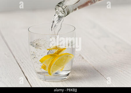 Das Gießen von Wasser aus einer Flasche in ein Glas mit Zitronenscheiben. Weiß Holz Hintergrund. Stockfoto