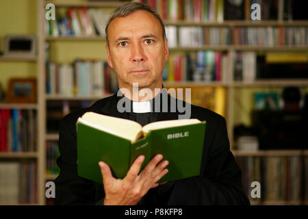 Eine gut aussehende ernsthafte katholische Priester ist das Studium, das Lesen der Bibel, in seine Bibliothek. Er schaut uns an. Stockfoto