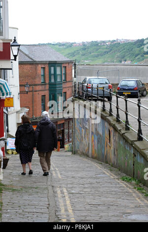 Ältere Paare zu Fuß den steilen Drop von Mildem Cliff, Scarborough, Großbritannien hinter den Geschäften und Street Art zum Meer in der Ferne Stockfoto