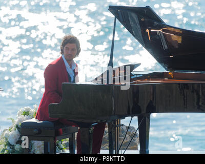 Newquay, Cornwall, England. 12. Juli 2018. Rosamunde Pilcher Schauspieler Manuel Mairhoffer spielt Klavier auf den Klippen, Filmen 'Meine Brüder Braut' Fistral Beach, UK, 12., Juli, 2018 Robert Taylor/Alamy Leben Nachrichten. Newquay, Cornwall, England. Credit: Robert Taylor/Alamy leben Nachrichten Stockfoto