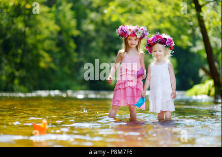 Zwei Entzückende kleine Schwestern tragen Blume Kronen spielen mit Papier Boote durch einen Fluss an warmen und sonnigen Sommer Tag Stockfoto