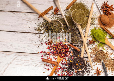 Von oben der Löffel aus Holz und Metall von unterschiedlichen Formen mit farbigen verschüttete Gewürze und Tees auf weissem Holztisch Stockfoto