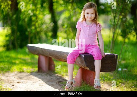 Adorable kleine Mädchen sitzen auf einer Bank in einem Park an einem warmen und sonnigen Sommertag Stockfoto