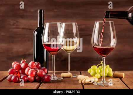 Rotwein wird in ein Glas von einer Flasche gegossen, auf einem dunklen Hintergrund, mit Kopie Raum und andere Gläser und Trauben im verschwommenen Hintergrund. Design Stockfoto