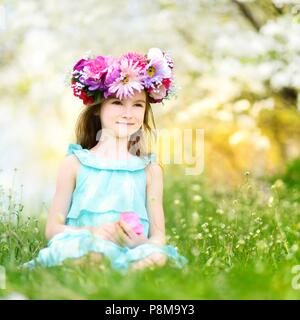 Adorable kleine Mädchen mit Blume Krone in Kirschblüten Garten auf schönen Frühlingstag sitzen Stockfoto