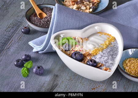 Joghurt mit Müsli, Blaubeeren, Mohn, Minze und zerdrückte Cornflakes im Herzen gedient - geformte Schale auf rustikalen Holztisch. Gesundes Frühstück anhand von quantitativen Simulatio Stockfoto