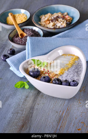 Joghurt mit Müsli, Blaubeeren, Mohn, Minze und zerdrückte Cornflakes im Herzen gedient - geformte Schale auf rustikalen Holztisch. Gesundes Frühstück anhand von quantitativen Simulatio Stockfoto