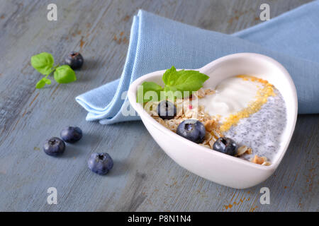 Joghurt mit Müsli, Blaubeeren, Mohn, Minze und zerdrückte Cornflakes im Herzen gedient - geformte bown auf rustikalen Holztisch. Gesundes Frühstück anhand von quantitativen Simulatio Stockfoto