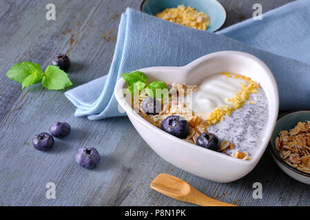 Joghurt mit Müsli, Blaubeeren, Minze und zerdrückte Cornflakes im Herzen gedient - geformte bown auf rustikalen Holztisch. Gesundes Frühstück Konzept. Stockfoto