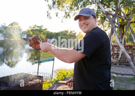 Grillfest in der Nähe von See oder Fluss im Freien. Man Grillen von Fleisch Rindfleisch Steak auf offenem Feuer brazier. Sommerurlaub und BBQ. Stockfoto