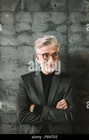 Vertikale Portrait von Happy reife Geschäftsmann stehend mit Waffen gegen einen grauen Hintergrund gekreuzt. Älterer Mann in formelle Kleidung und SMI Stockfoto