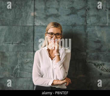 Porträt der schönen jungen Frau in formale Abnutzung gegen graue Wand stehend und lächelnd. Geschäftsfrau mit Brille mit Kamera suchen mit Hand auf Chi Stockfoto