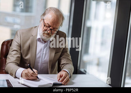 Alten Professor mit grayhair und Bart im schönen Anzug Notizen in das Notebook im Zimmer mit Panorama Fenster Stockfoto