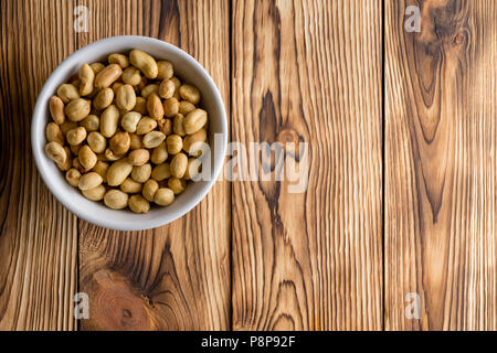 Generische weiße Schüssel voller Frisch geröstete Erdnüsse ohne Schale in der Ecke auf einem rustikalen Holztisch mit Kopie Raum von oben gesehen Stockfoto
