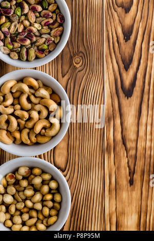 Drei kleine weiße Generische der sortierten Nüsse mit Cashewnüssen, Pistazien und Erdnüsse auf einem rustikalen Holztisch mit Copyspace arrangiert als seitliche vertikale Bord Stockfoto