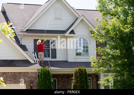 Mann äußere Reinigung der oberen Etage seines Hauses stehend auf das untere Dach mit Hilfe einer Druck Sprüher das Holz Verschalungen und Friese an den zu waschen. Stockfoto