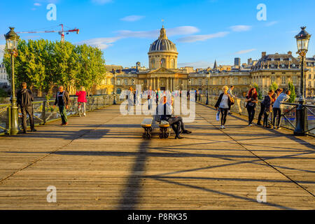 Paris, Frankreich, 1. Juli 2017: Touristen sitzen auf Bänken von Pont des Arts bei Sonnenuntergang, bei Fußgänger-Brücke, die über den Fluss Seine. Institut de France mit Bibliotheque Nazarine für den Hintergrund. Stockfoto