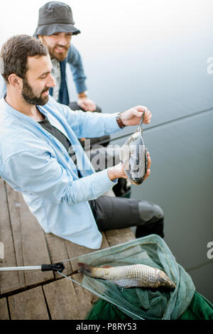 Zwei glückliche Fischer holding Fisch sitzen auf dem Holzsteg beim Fischen auf dem See am Morgen Stockfoto