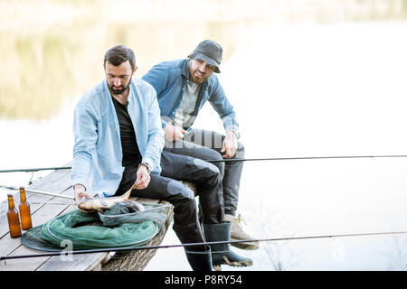 Zwei glückliche Fischer holding Fisch sitzen auf dem Holzsteg beim Fischen auf dem See am Morgen Stockfoto