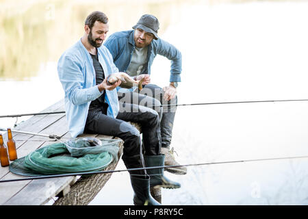 Zwei glückliche Fischer holding Fisch sitzen auf dem Holzsteg beim Fischen auf dem See am Morgen Stockfoto