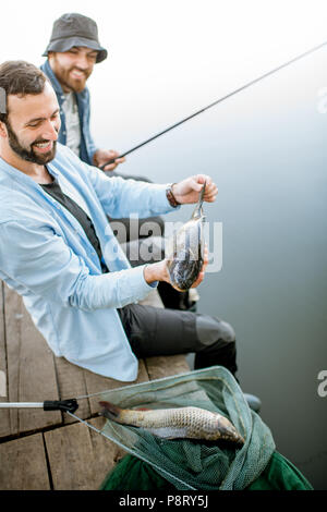 Zwei glückliche Fischer holding Fisch sitzen auf dem Holzsteg beim Fischen auf dem See am Morgen Stockfoto